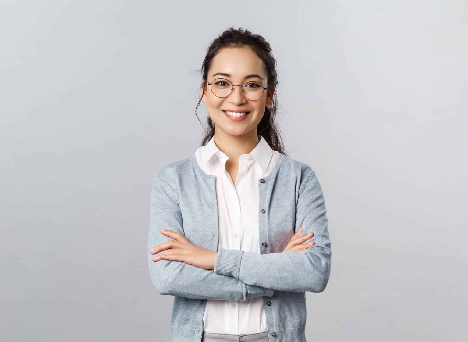 Mujer joven con características similares a la de la imagen anterior, con gafas, camisa blanca y cárdigan azul claro. Está sonriendo con los brazos cruzados. El fondo es gris claro.